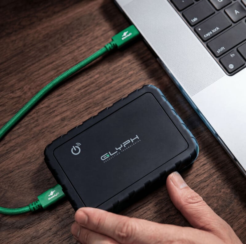 A person’s hand holding a portable Glyph external hard drive connected to a laptop with a green USB cable on a wooden desk.