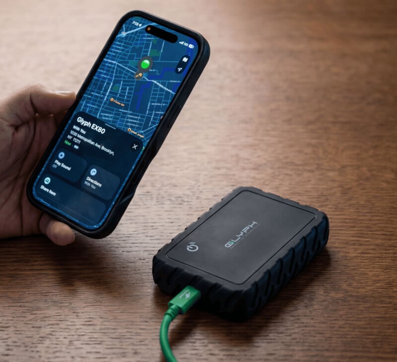 A hand holds a smartphone displaying a map and device info, next to a rugged external hard drive connected by a green cable, both placed on a wooden surface.