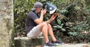 A man in a cap and shorts sits on a stone ledge in a forest, holding a butterfly on one hand and photographing it up close with a camera in the other hand. Lush green plants surround him.