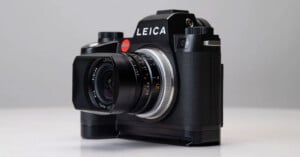 A black Leica camera with a large lens and textured grip sits on a light surface against a plain, light gray background. The brand name "Leica" is prominently displayed above the lens.