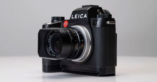 A black Leica camera with a large lens and textured grip sits on a light surface against a plain, light gray background. The brand name "Leica" is prominently displayed above the lens.