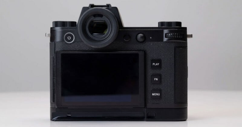 A close-up view of the back of a black digital camera showing its screen, viewfinder, control buttons, and textured grip, placed on a white surface with a plain background.