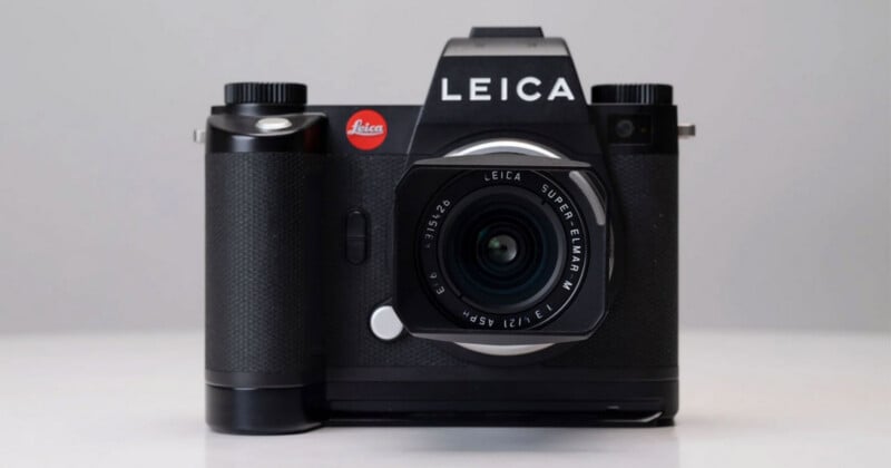 A black Leica digital camera with a prominent lens, displayed on a white surface against a plain light gray background. The camera has the Leica logo and branding visible on the body.