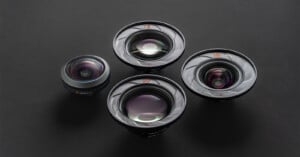 Four camera lenses of varying sizes are arranged on a dark surface. The lenses are labeled 21, 60, 75, and 100, with visible reflections on their glass elements.