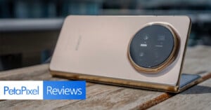 A gold-colored smartphone with a large circular camera module is propped up on a wooden surface. The “PetaPixel Reviews” logo appears in the bottom left corner of the image.