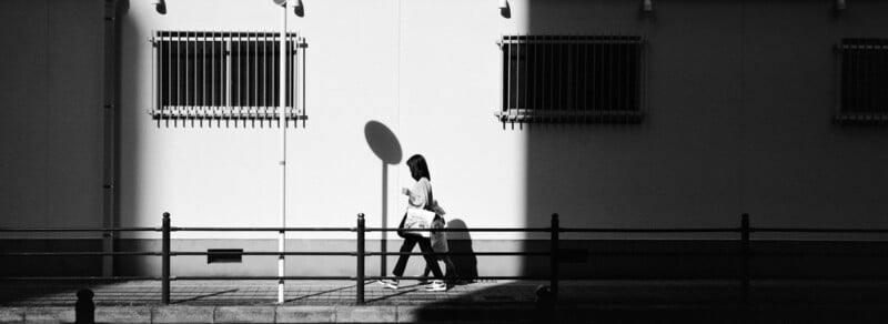 A person walks along a sidewalk in bright sunlight, casting a long shadow on a white wall with barred windows. The scene is in black and white and features strong contrasts between light and dark.