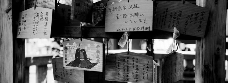 Wooden ema plaques with handwritten Japanese messages hang on display at a shrine. The black and white image highlights the details of the plaques and the surrounding wooden structure.