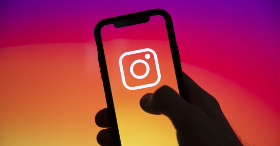 A hand holds a smartphone displaying the white Instagram logo on a screen with a vibrant pink, orange, and yellow gradient background.