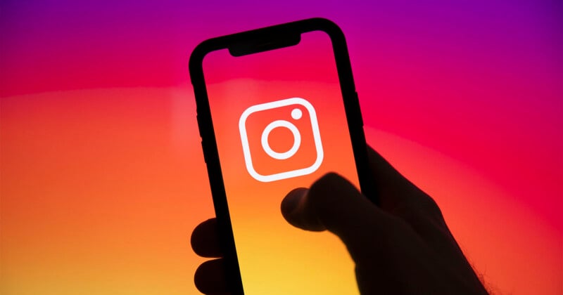 A hand holds a smartphone displaying the white Instagram logo on a screen with a vibrant pink, orange, and yellow gradient background.