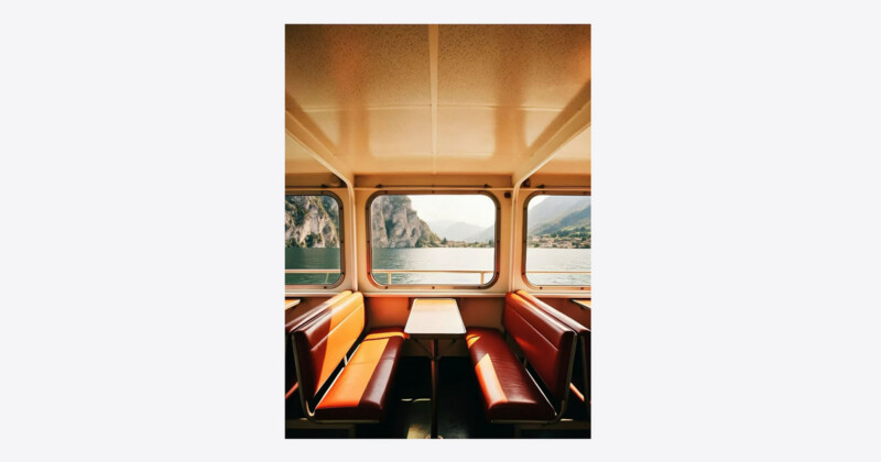 Sunlight streams into a ferry cabin with empty red seats and a small table, offering a view through large windows of a lake, distant mountains, and rocky cliffs under a clear sky.