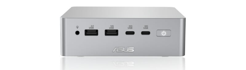 A silver ASUS mini PC showing the front panel, which includes a headphone jack, two USB-A ports, two USB-C ports, and a power button. The ASUS logo is visible at the bottom.