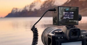 A professional camera with an LRTimelapse PRO Timer attached is set up by a calm lake at sunset, with snow-capped mountains and a colorful sky reflected in the water.