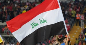 A person waves a large Iraqi flag with red, white, and black horizontal stripes and green Arabic script at a sports stadium with a cheering crowd in the background.