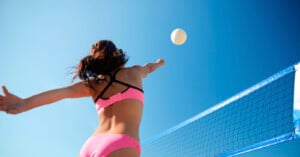 A woman in a pink bikini jumps to hit a volleyball over a blue net on a sunny day, set against a clear blue sky.