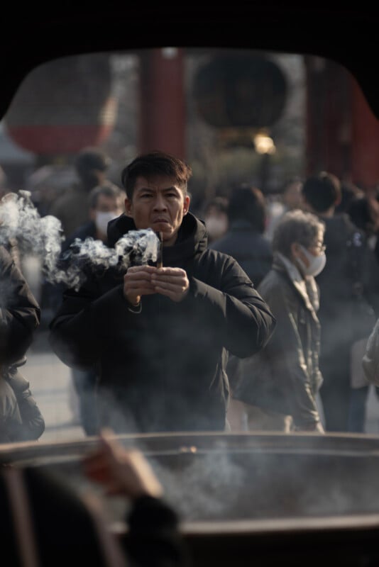 A man in a black jacket stands among a crowd, holding his hands over rising smoke, with people in the background, some wearing masks. The scene appears to be outdoors and has a solemn atmosphere.