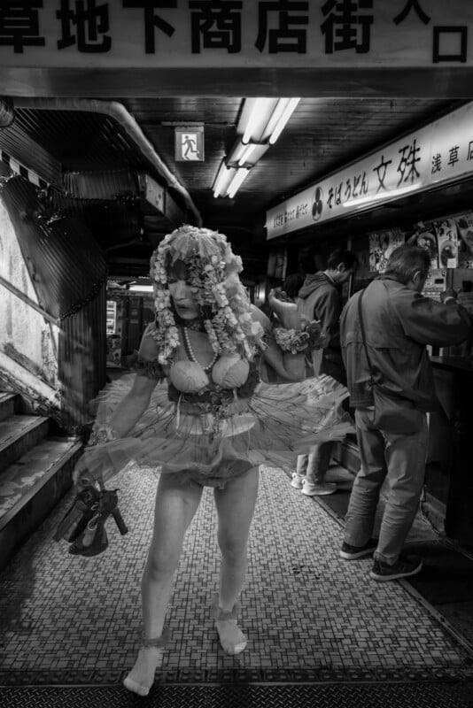 A person in an elaborate costume with floral headpieces and baloons performs in an underground shopping area, while others stand nearby at vending machines; the scene is in black and white.
