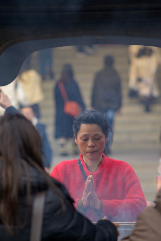 A woman in a bright red sweater stands with her hands pressed together in prayer, eyes closed, surrounded by smoke, with blurred people and stone steps in the background.