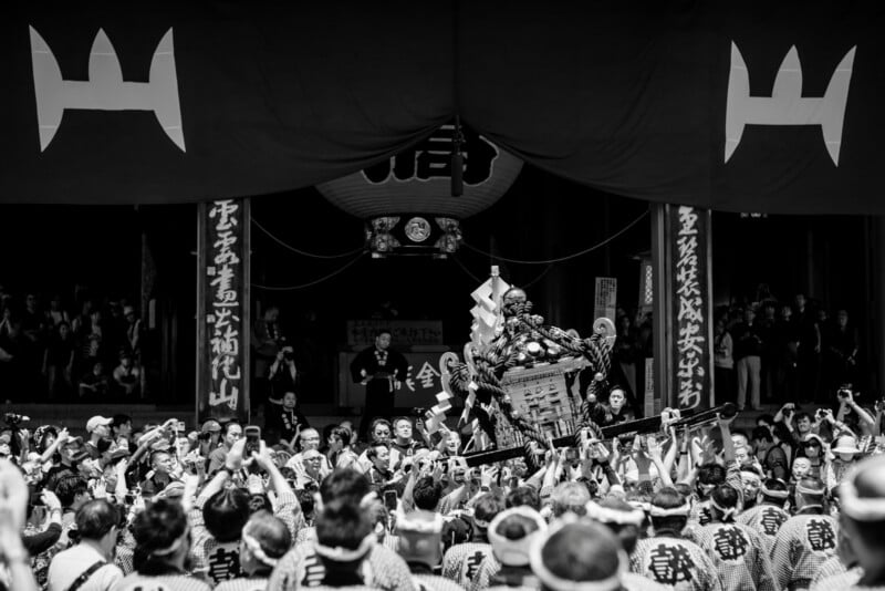 A large crowd in traditional clothing carries an ornate mikoshi shrine during a Japanese festival, with banners and lanterns decorating the temple entrance in the background. The photo is in black and white.