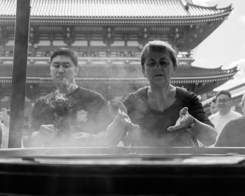 Two people stand in front of a large incense burner at a temple, with smoke rising around them as they wave their hands. A traditional building with a tiled roof is visible in the background. The image is in black and white.