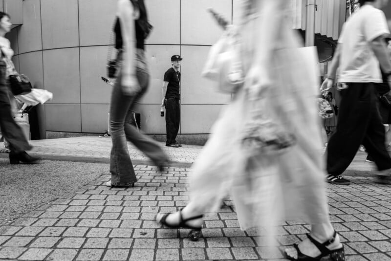 Black and white photo of a city street with blurred pedestrians walking, capturing a sense of motion. A man in a dark shirt and cap stands still against a tiled wall, watching the passing crowd.