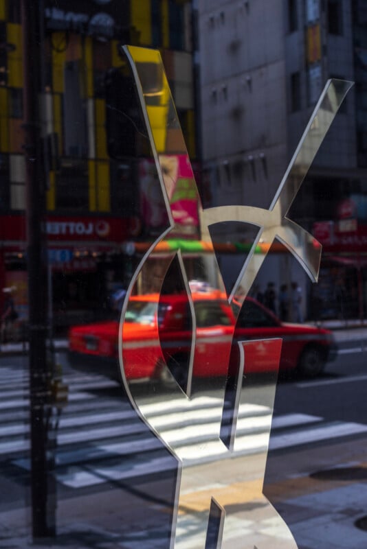 A red car drives past a city street, reflected in a shop window featuring a large, metallic Yves Saint Laurent (YSL) logo. Buildings and a crosswalk are visible in the background.