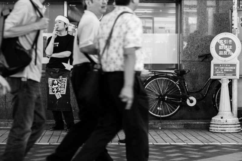 A black-and-white street scene shows people walking past a man standing outside a building, wearing an apron and headband. A bicycle is parked against the wall, and there is a sign with Japanese text near a lamppost.