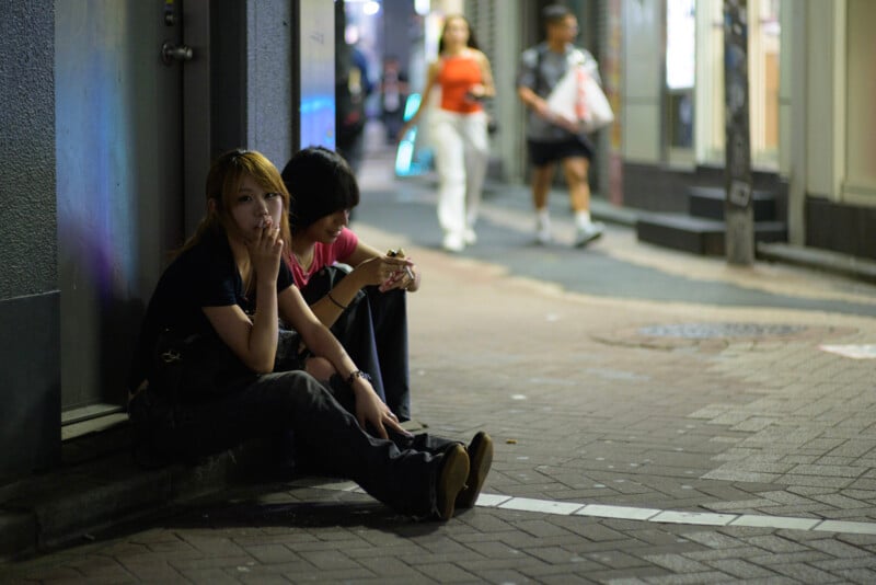 Two people sit on a city sidewalk at night, one woman smoking a cigarette and looking at the camera. Blurred pedestrians walk by on the lit street in the background. The scene feels urban and casual.