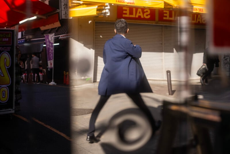 A man in a blue coat walks energetically across a sunlit street, with "SALE" signs and closed shop shutters in the background and blurred objects creating reflections in the foreground.