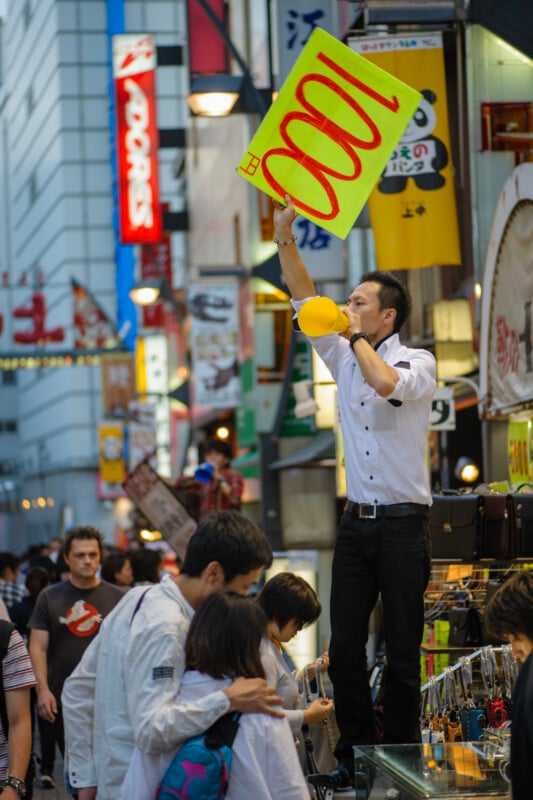 A man stands on a platform in a busy street, holding a large yellow sign with “1000” written on it and blowing a yellow horn. Crowds of people walk by, surrounded by colorful signs and shopfronts.