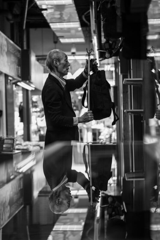 An elderly man in a suit examines a backpack in a shop, his reflection visible on a glossy surface below him. The image is in black and white, creating a contemplative and quiet atmosphere.