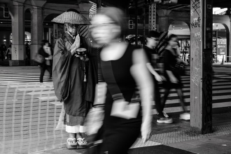 A monk in traditional robes and a straw hat stands with hands pressed together on a busy street, while blurred pedestrians move past in the foreground, conveying motion and stillness.