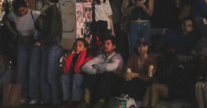 A group of people, some standing and some sitting, watch something off-camera at night. Several hold drinks, and the scene is dimly lit. A wall with posters is visible in the background.