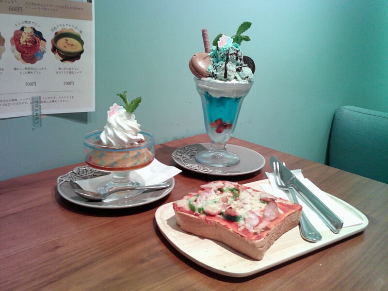 A table with a slice of pizza toast, a glass parfait with blue jelly and whipped cream, and a dessert topped with whipped cream and a mint leaf. A fork and knife are placed nearby. Japanese menu on the wall in the background.