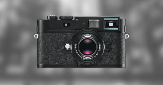 A black Leica digital camera with a textured grip, prominent lens, and various dials is centered against a blurred gray background.