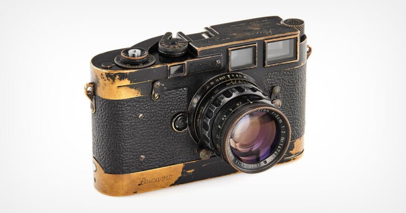 A vintage black and gold Leica camera with visible signs of wear, including scratched and faded paint on the edges, sits on a plain white background.