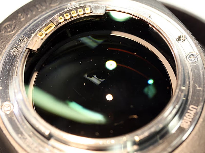 Close-up of a camera lens, showing reflections on the glass, the metal mount, and electronic contacts around the rim. Dust particles and light glares are visible on the lens surface.