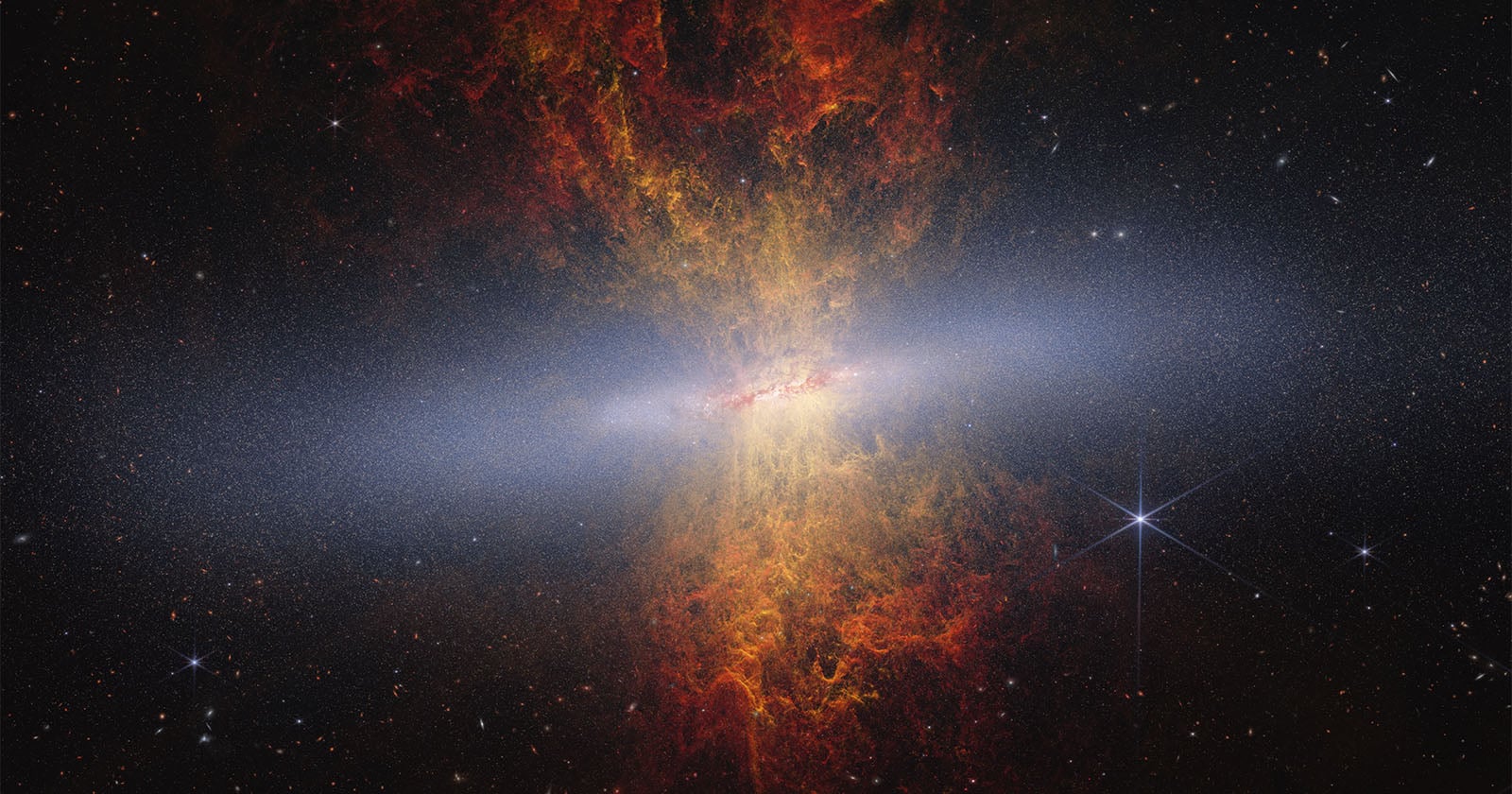 A vibrant galaxy with a bright, glowing center emits orange-red clouds of gas outward, forming a fiery X-shape against a backdrop of stars and cosmic dust in deep space.