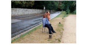 A person wearing a horse mask, blue shirt, and black pants sits on a folding chair beside a wet road and a gravel path, with trees and a stone wall in the background.