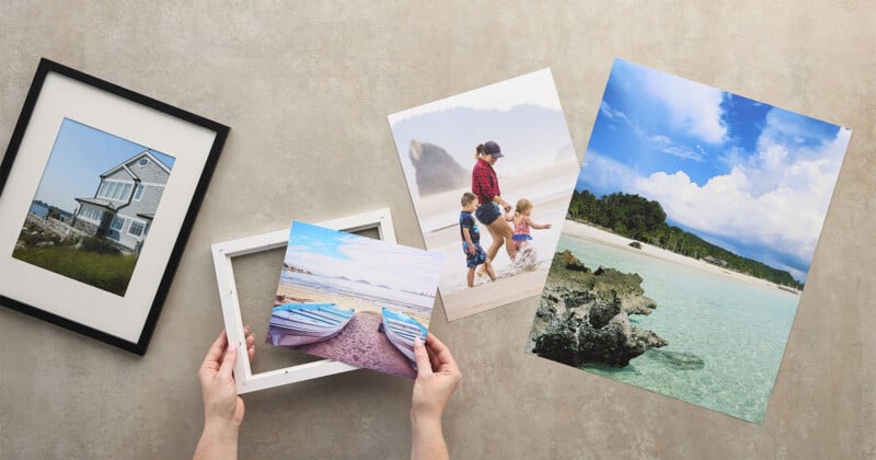 A person frames a beach photo, surrounded by prints of a house, a family on the beach, and a tropical seascape, all laid out on a neutral surface.