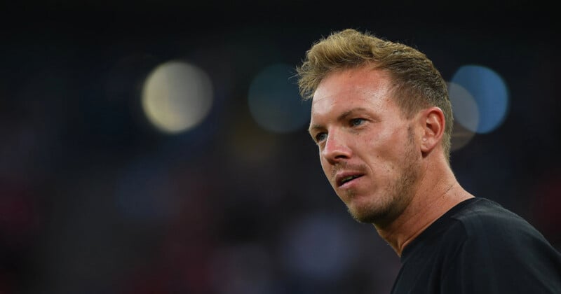 A man with short blond hair and light facial stubble looks to the side, wearing a black shirt, with a blurred dark background and circular lights behind him.