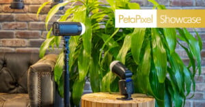 A studio light stands on a tripod next to a black camera flash on a wooden stump, with a large green plant and a brown leather chair in the background. The text "PetaPixel Showcase" appears in the top right corner.