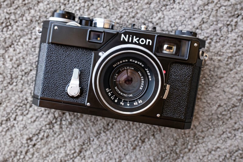 A vintage black Nikon rangefinder camera with a large lens is resting on a textured gray carpet. The camera has dials and buttons on top, and "Nikon" is written above the lens.