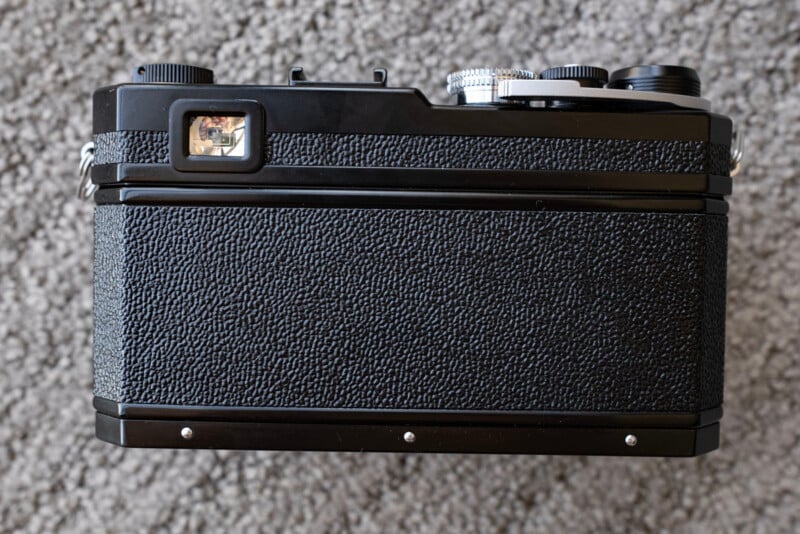 The image shows the back view of a black film camera with a textured grip, a small rectangular viewfinder on the left, and control dials on the top, placed on a gray carpeted surface.