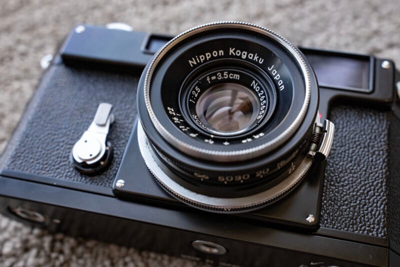 Close-up of a vintage black camera with a Nippon Kogaku Japan 5cm f/3.5 lens, resting on a textured grey surface. The lens details and camera body are clearly visible.