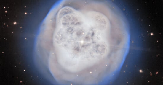 A glowing, cloud-like planetary nebula with a bright center surrounded by soft blue and white halos, set against a star-filled black background.