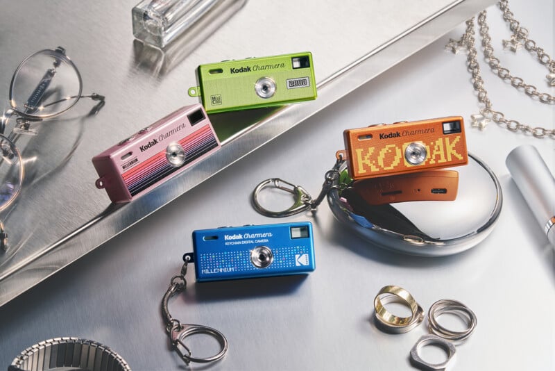 Four colorful Kodak Charman mini cameras on a metallic surface, surrounded by rings, a chain, eyeglasses, a lipstick, and a perfume bottle, arranged in a stylish, organized layout.
