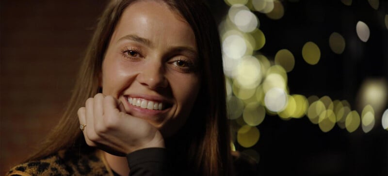 A woman with long brown hair smiles warmly at the camera, resting her chin on her hand. She wears a ring and dark clothing. The background features soft, out-of-focus lights, creating a cozy atmosphere.