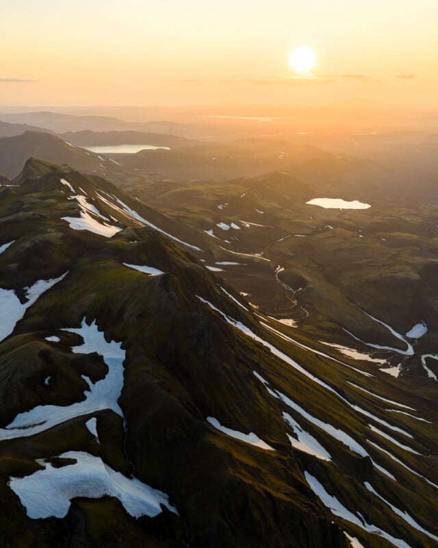 Aerial view of a mountain ridge with patches of snow, green valleys, winding rivers, and small lakes at sunset, under a golden sky.