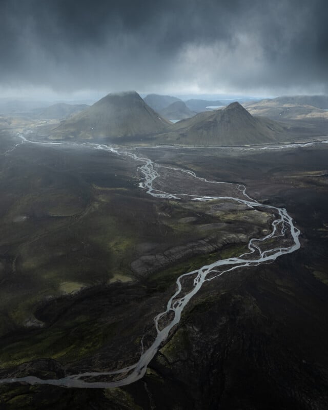 A winding river flows through a dark, rugged landscape with scattered green patches, leading toward distant mountains under a dramatic, cloudy sky.