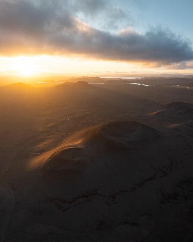 Aerial view of volcanic craters at sunrise, with soft sunlight casting golden hues over the landscape and distant clouds partially covering the sky.